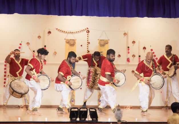 Nine Tamil musicians performing on stage with drums, wearing red, gold and white costumes.
